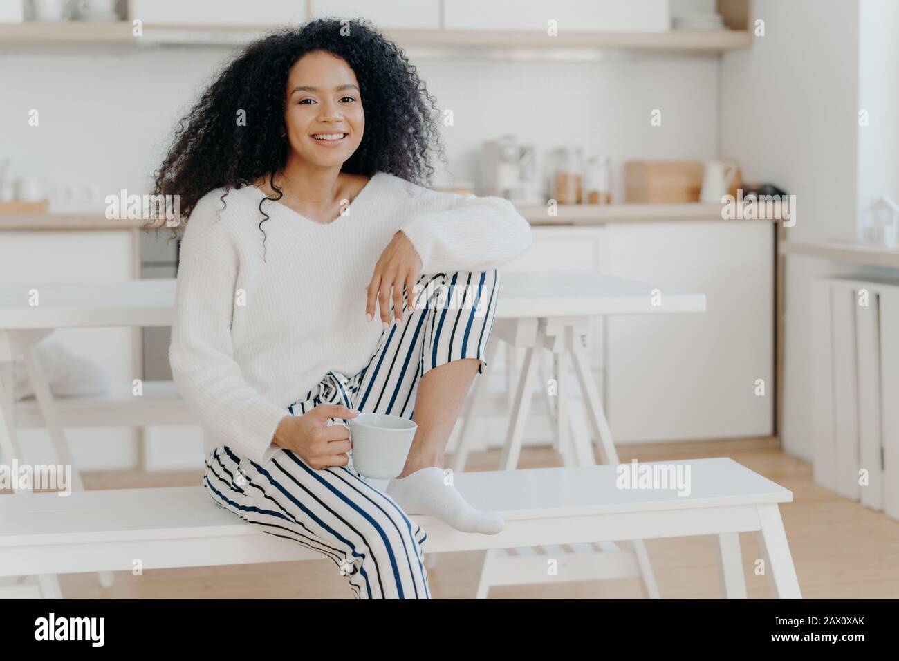 Glad African American woman holds cup of hot beverage, leans at knee, wears white stylish jumper and striped trousers, smiles pleasantly, spends leisu Stock Photo