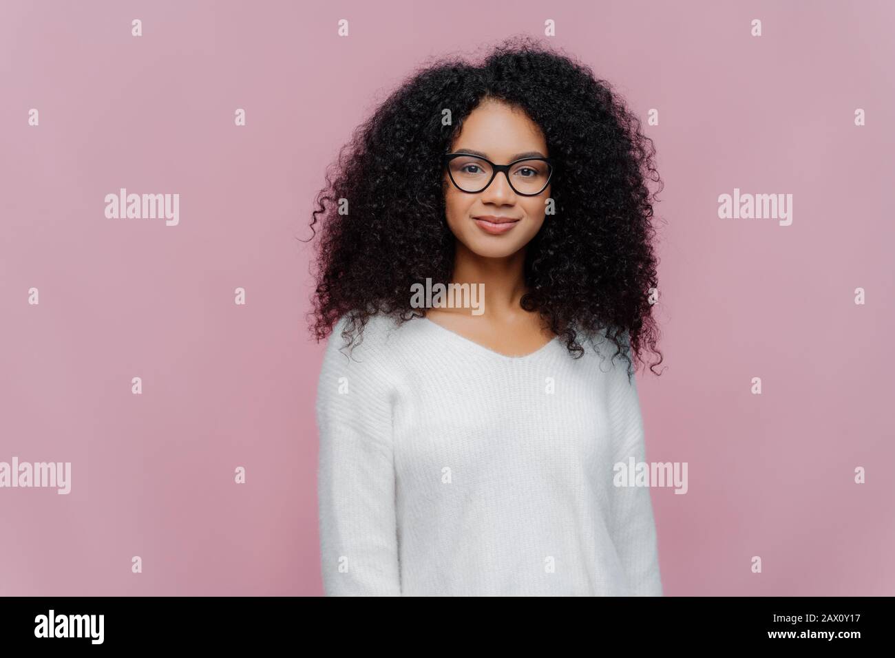 Half length shot of attractive African American woman looks through transparent glasses, white sweater, has serious calm expression, poses against vio Stock Photo