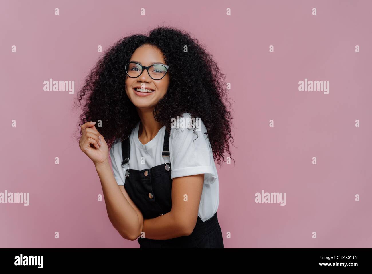 Studio shot of happy dark skinned lady with crisp hair, has dreamy expression, wears optical glasses, stylish clothes, imagines something pleasant, st Stock Photo