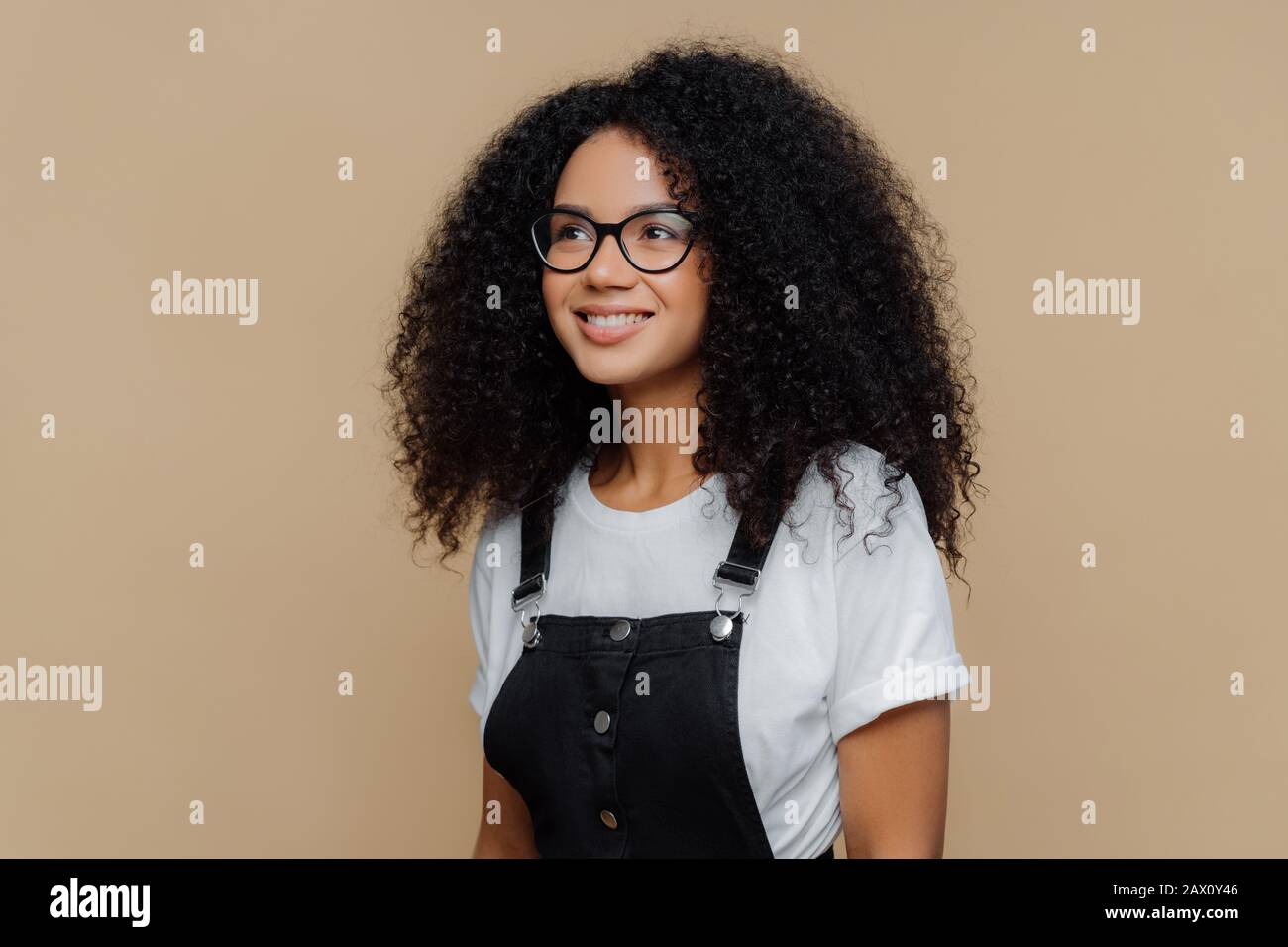 Pleased dreamy woman with curly hairstyle, looks through transparent glasses,  dressed in casual white t shirt and black dungarees, poses against brown  Stock Photo - Alamy