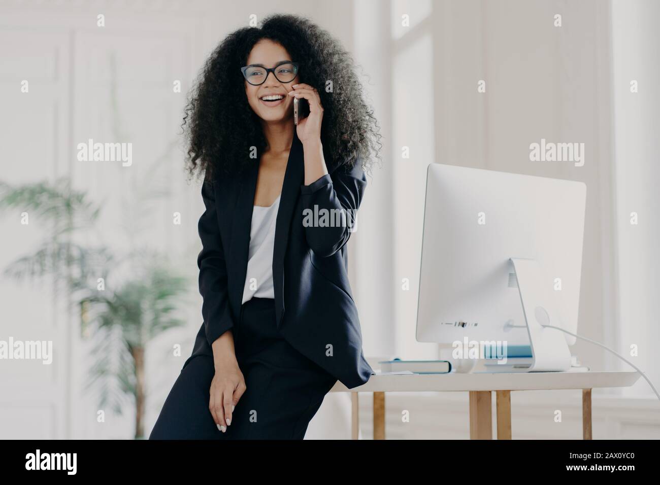Image of satisfied dark skinned businesswoman leans at white table with computer, has break after work, poses in her cabinet, speaks via cellular, wea Stock Photo