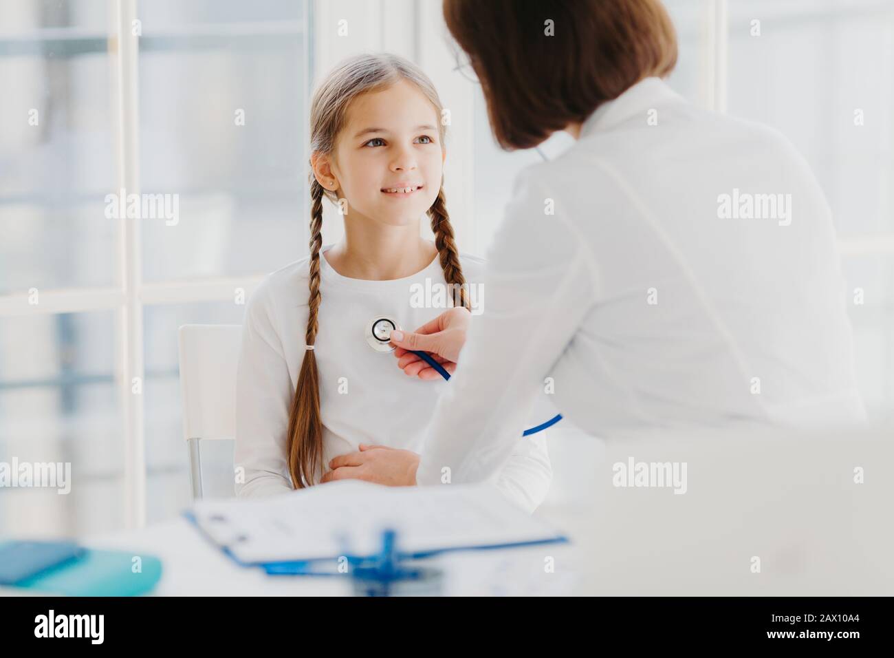 Family general practitioner examines child, listens lungs with stethoscope, makes prescription pose in moren hospital office. Ill kid has cold, comes Stock Photo