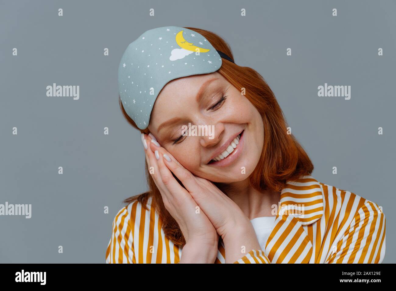Close up shot of lovely tender woman has ginger hair, minimal makeup, leans at palms pressed together, smiles positively, wears blindfold on head, see Stock Photo
