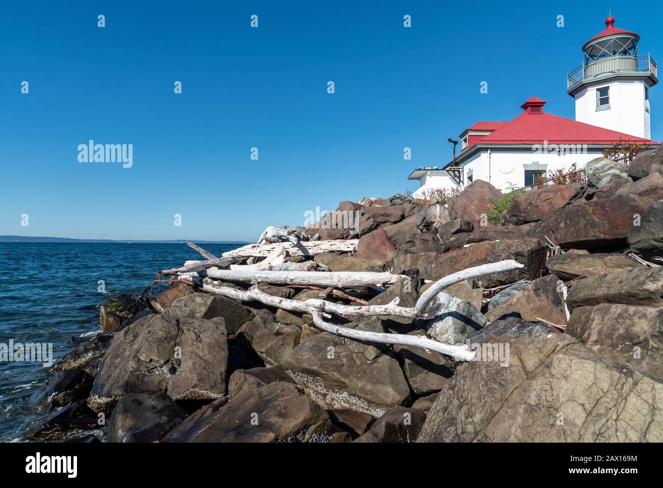 Sun bleached driftwood resting on reinforced rock sea wall under the Alki Point Lighthouse in West Seattle, Washington, mid day blue sky. Stock Photo