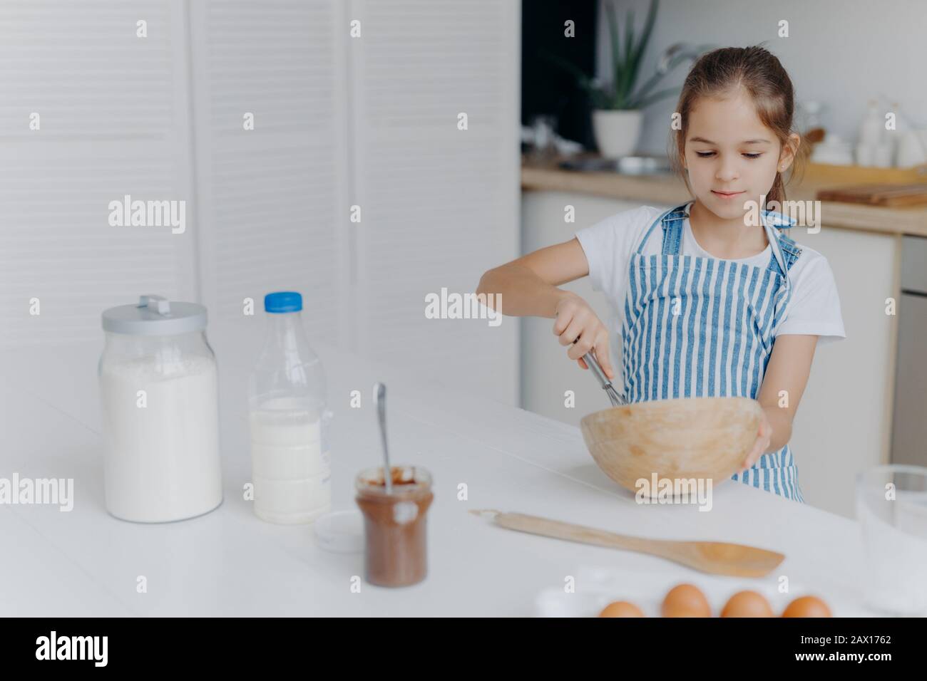 Children, cooking and home concept. Pretty dark haired girl whisks ingredients in bowl, busy preparing dough for cake, being future chef, wears white Stock Photo