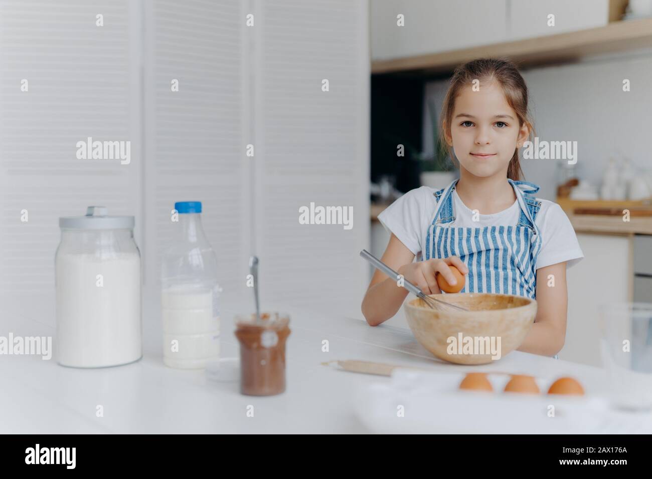 Adorable European child enjoys cooking activity, breaks egg in bowl, whisks ingredients, prepares tasty pastry, being young cook, wears apron, poses a Stock Photo
