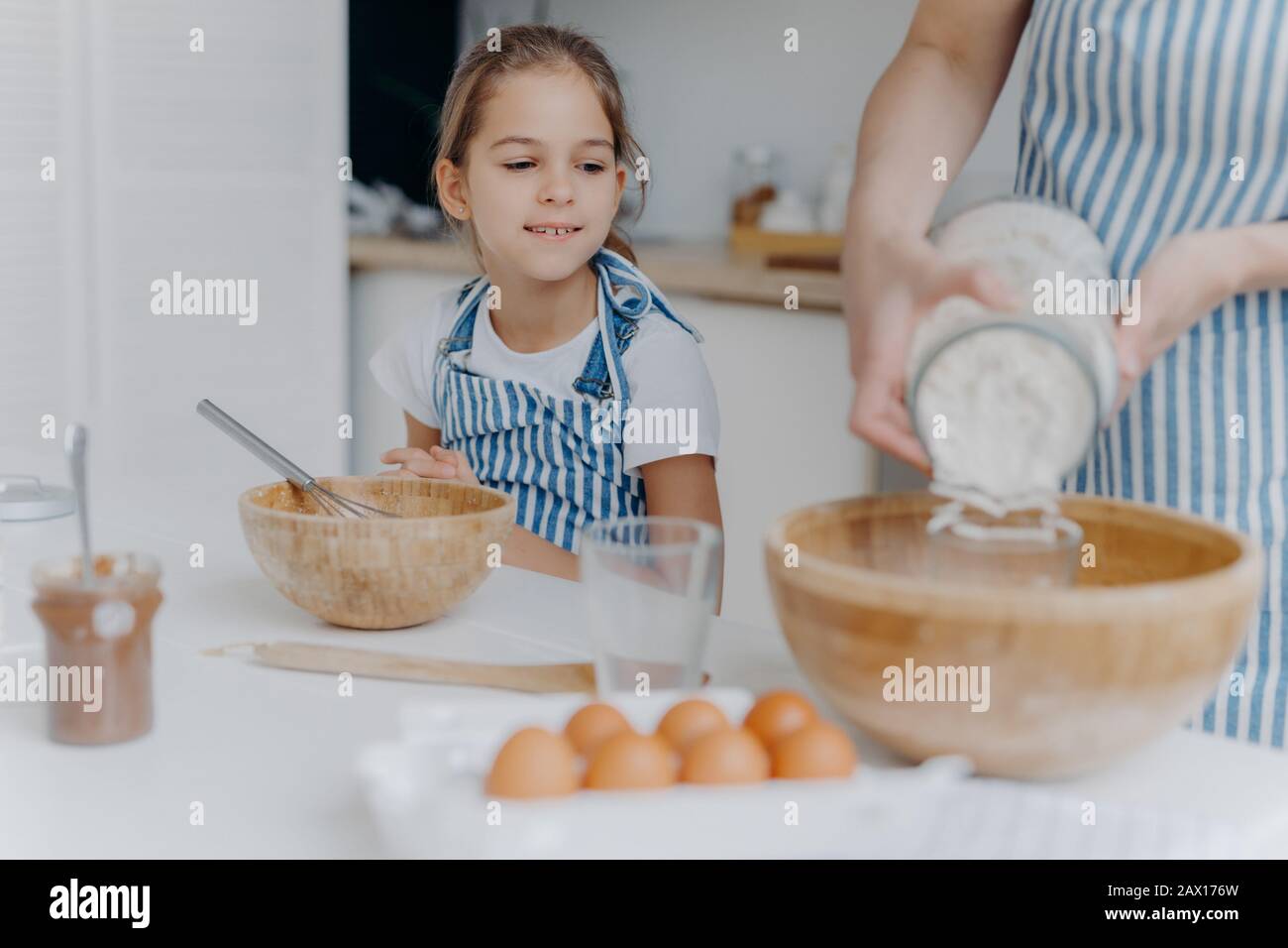 Curious little girl looks how mom prepares dough for pastry, learns to cook, gets culinary experience, wears apron. Faceless woman adds flour in bowl Stock Photo