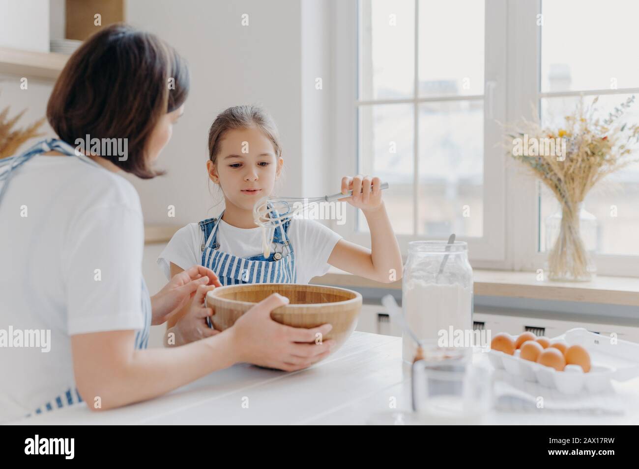 Adorable little child in apron shows whisk with white cream, wears apron, cooks together with mommy, pose in spacious kitchen together, make cake for Stock Photo
