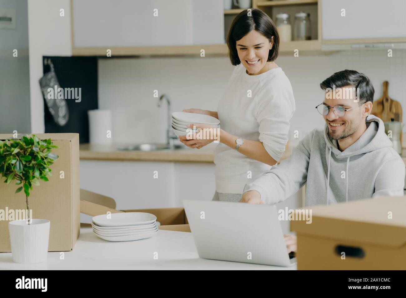 Moving house and real estate. Caring brunette woman holds pile of plates, unpacks personal belongings, husband asks her advice, chooses something to b Stock Photo