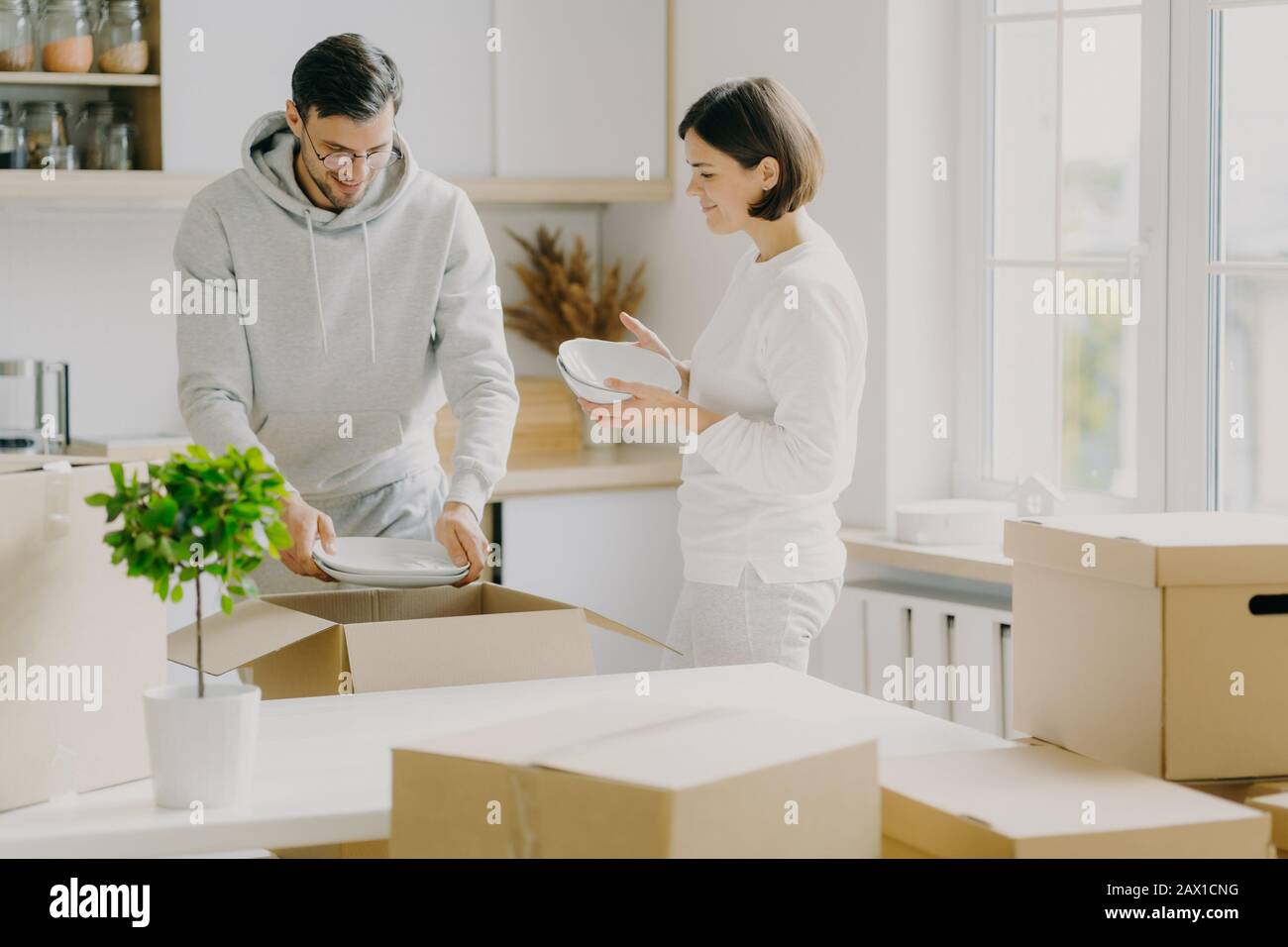 Photo of glad married woman and man unpack dinnerware from boxes, dressed casually, hold plates, pose in new modern kitchen, surrounded with moving pa Stock Photo