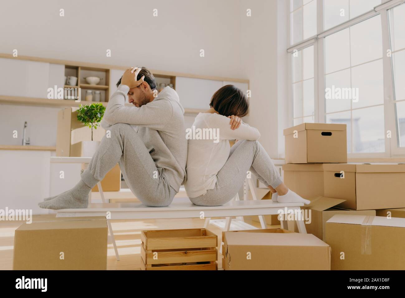 Photo of stressful couple get divorse, leave house, start living seperately, sit backs to each other, pose in kitchen, cardboard boxes with belongings Stock Photo