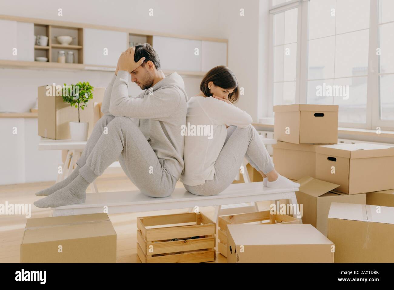 Unhappy young married woman and man have to leave house, move in other place, sit back to each other pose in empty room with stack of boxes, wear dome Stock Photo