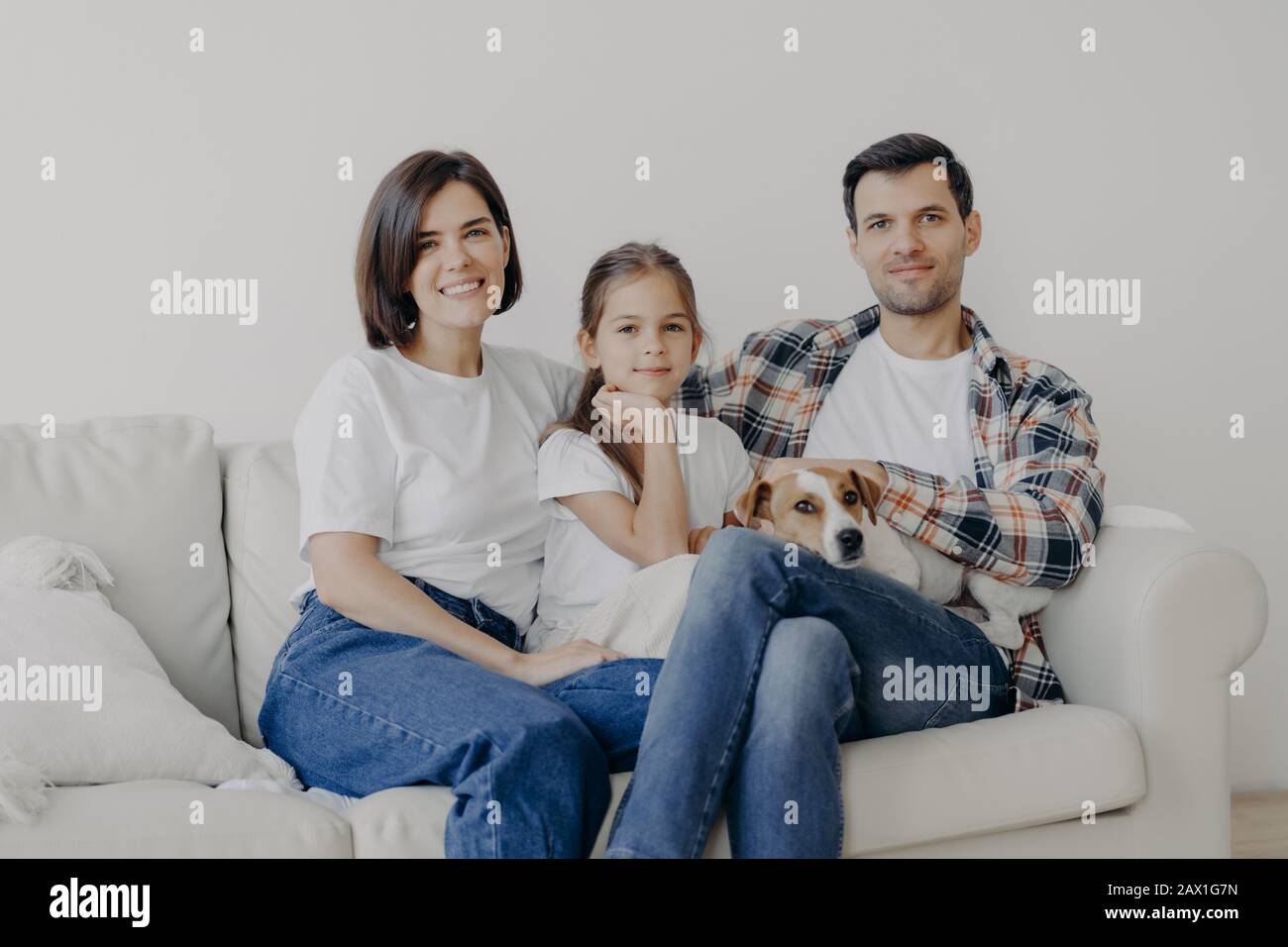 Photo of happy friendly family of mum, dad and daughter pose on comfortable sofa with pet, have good relationship, dressed casually, smile positively. Stock Photo