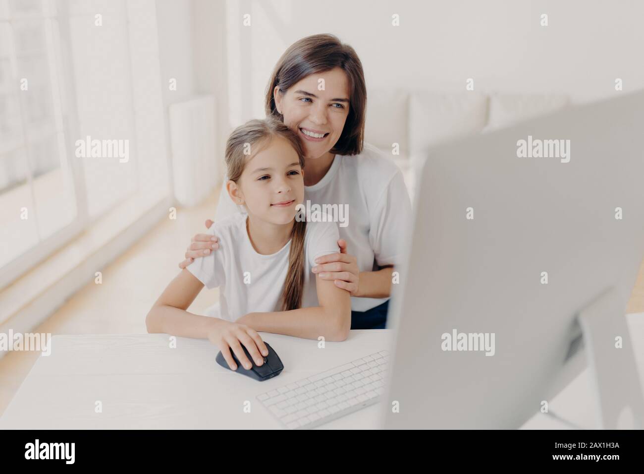 Happy affectionate mom embraces her small daughter, work together on modern computer, preapare presentation for lesson, look gladfully into monitor, s Stock Photo