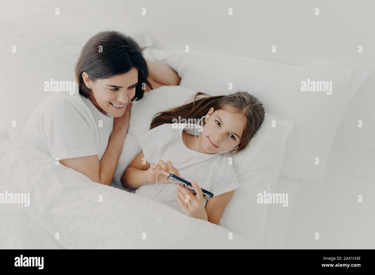 Caring mom looks attentively at her little daughter who wakes up early, listens music in earphones, searches new song in playlist of modern smartphone Stock Photo