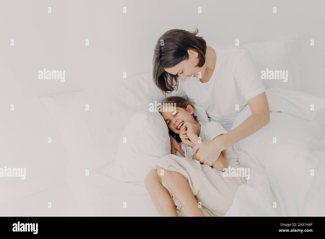 Top view of happy funny girl in pyjamas and her caring mom pose on bed, play together in bed, enjoy softness and comfort, express positive emotions. F Stock Photo