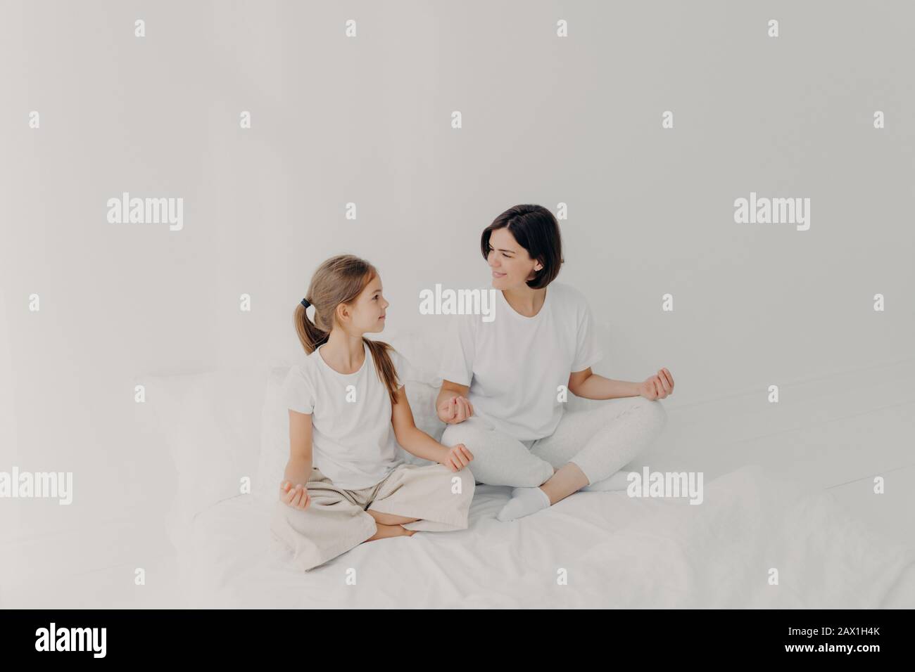 Indoor shot of happy mom and daughter have pleasant talk together, have yoga practice, meditate on soft bed, dressed casually, pose on white bedclothe Stock Photo