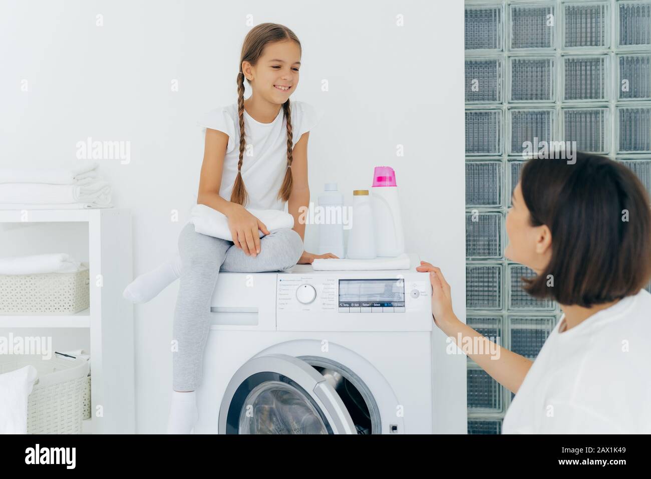 Adorable small girl with pigtails, poses on top of washer, holds white soft towel, looks gladfully at mother, talk about plans after washing. Brunette Stock Photo
