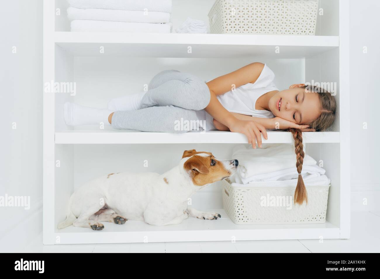 Sleepy little girl feels comfortable as lies on white shelf dressed in casual clothing tired after washing her dog looks attentively. White color. Kid Stock Photo