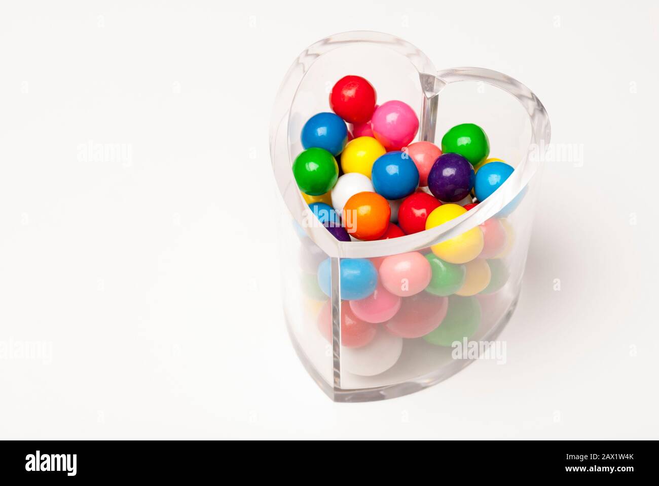 Clear heart-shaped candy jar filled with bright, colorful Valentine gumball candies on white background Stock Photo