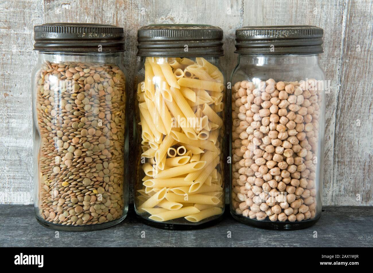 Vintage glass Kilner storage food jars with metal lids containing pasta, lentils and chickpeas against wooden background in kitchen.  Zero wastes concept Stock Photo
