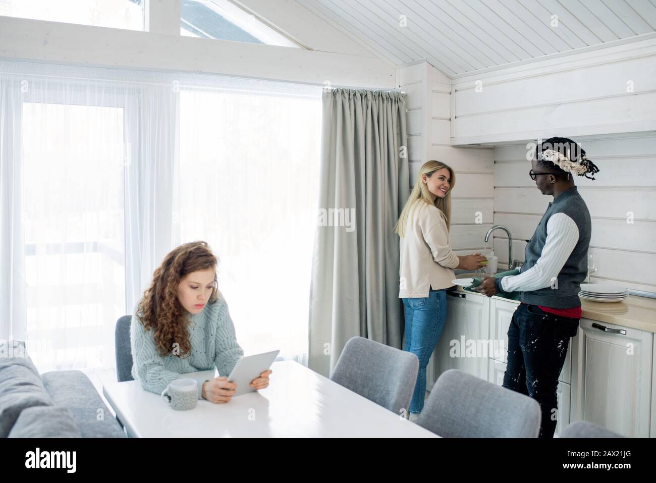 Blond woman chatting with Black guy while washing dishes, their friend sitting at table and reading something using tablet PC Stock Photo