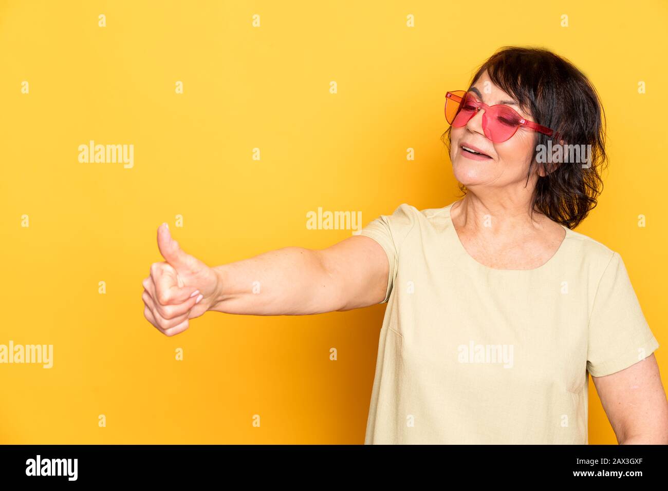 Beautiful Senior woman in pink neon sunglasses showing like by fingers over yellow background Stock Photo
