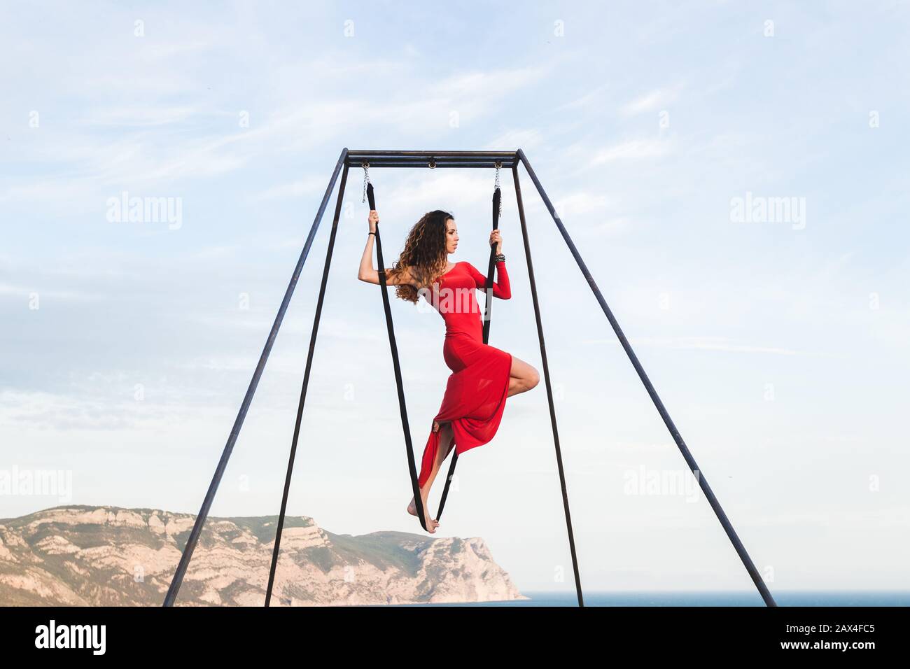 Woman in red dress practicing pole fly dance poses in a hammock outdoor with a mountain view. Female sports, wellbeing concept. Pilates outdoors. Stock Photo