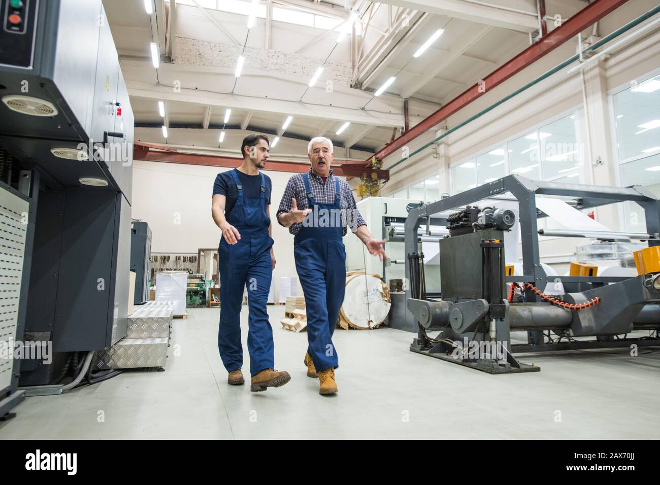 Experienced senior man in blue overall gesturing hands while interacting with young worker at printing factory Stock Photo