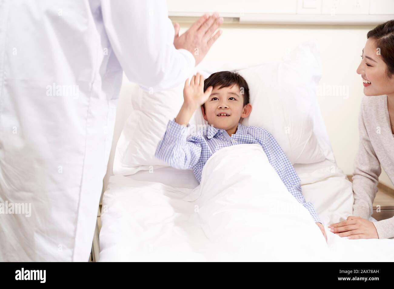 happy five year old asian kid lying in bed in hospital ward giving doctor a hi-five Stock Photo