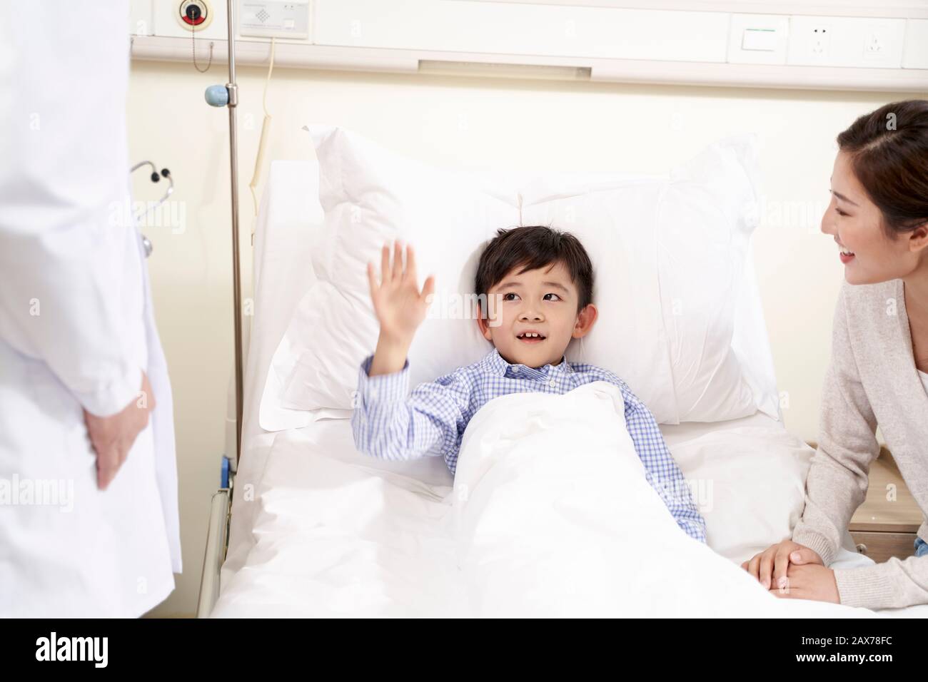 happy five year old asian kid lying in bed in hospital ward waving good-bye to doctor Stock Photo