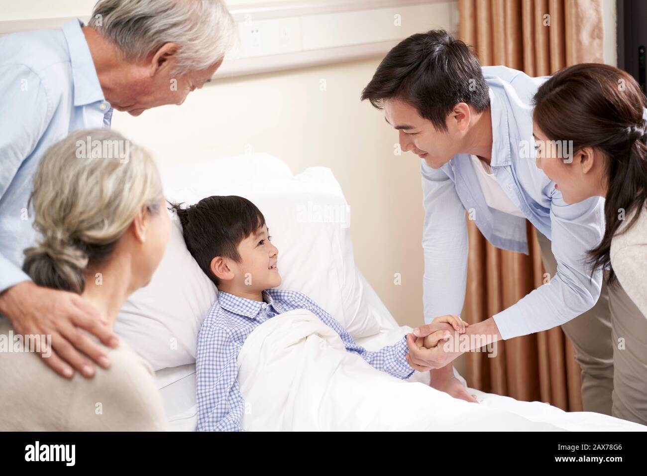 five year old asian child lying in bed in hospital ward gets a visit from parents and grandparents Stock Photo