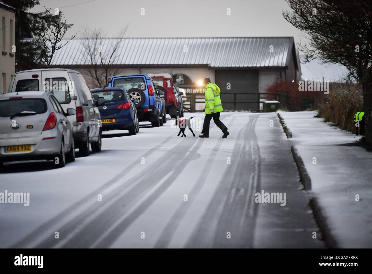 A dog walker crosses snow covered roads in Princetown on the top of Dartmoor, Devon, where snow has fallen on high ground. Stock Photo