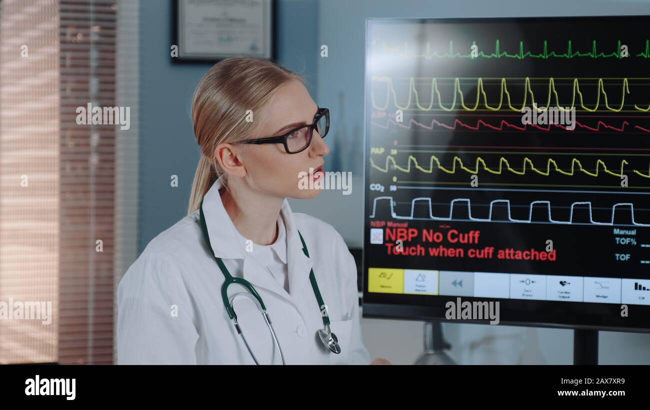 Close-up of beautiful female doctor in lab coat discussing EKG data with colleague showing on big display in modern sports lab. Stock Photo