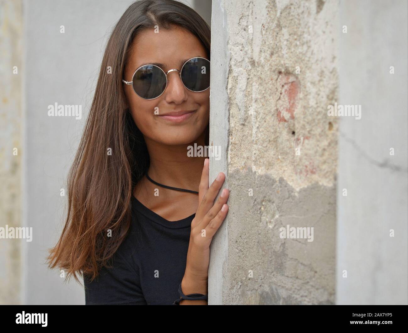 Young woman with beautiful hair and sunglasses looks around the corner of an old house. Stock Photo