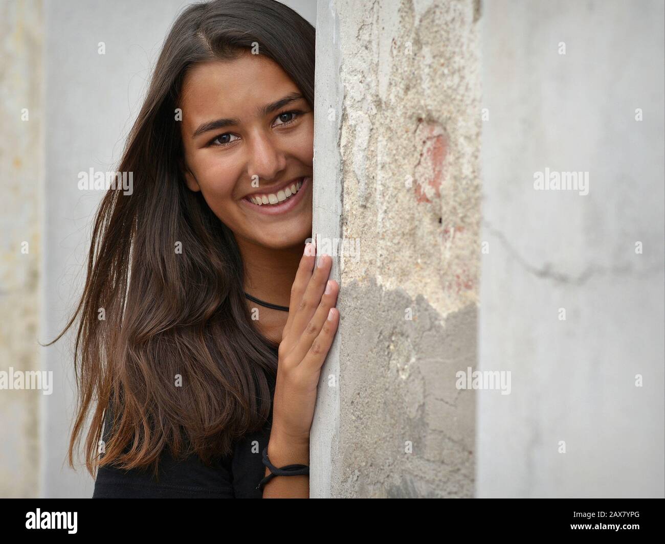 Young woman with beautiful hair smiles and looks around the corner of an old house. Stock Photo