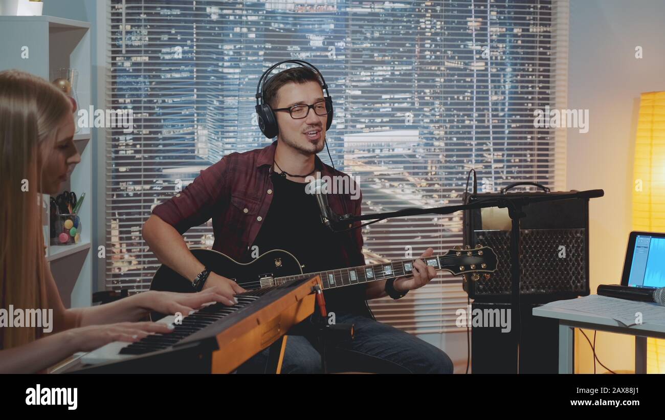 Recording hit song in home studio: young man in headphone singing and playing guitar while the pianist accompanying him. There is different recording equipment in the room. Stock Photo
