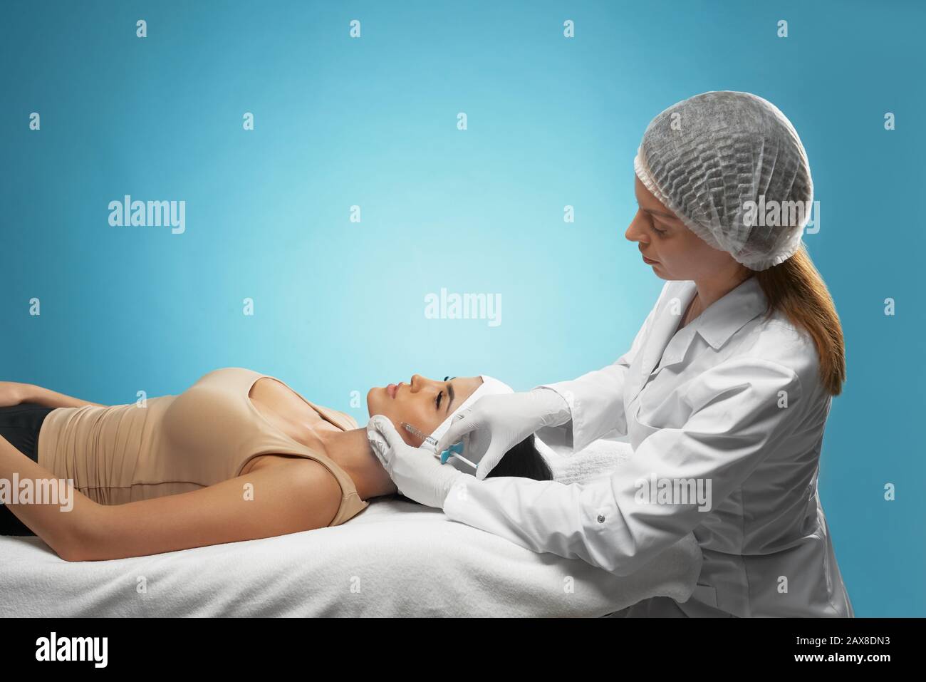 Side view of injection in female cheek. Cosmetologist in doctors coat using syringe with special liquid and cotton pad while brunette patient in towel on head lying. Concept of cosmetology, beauty. Stock Photo