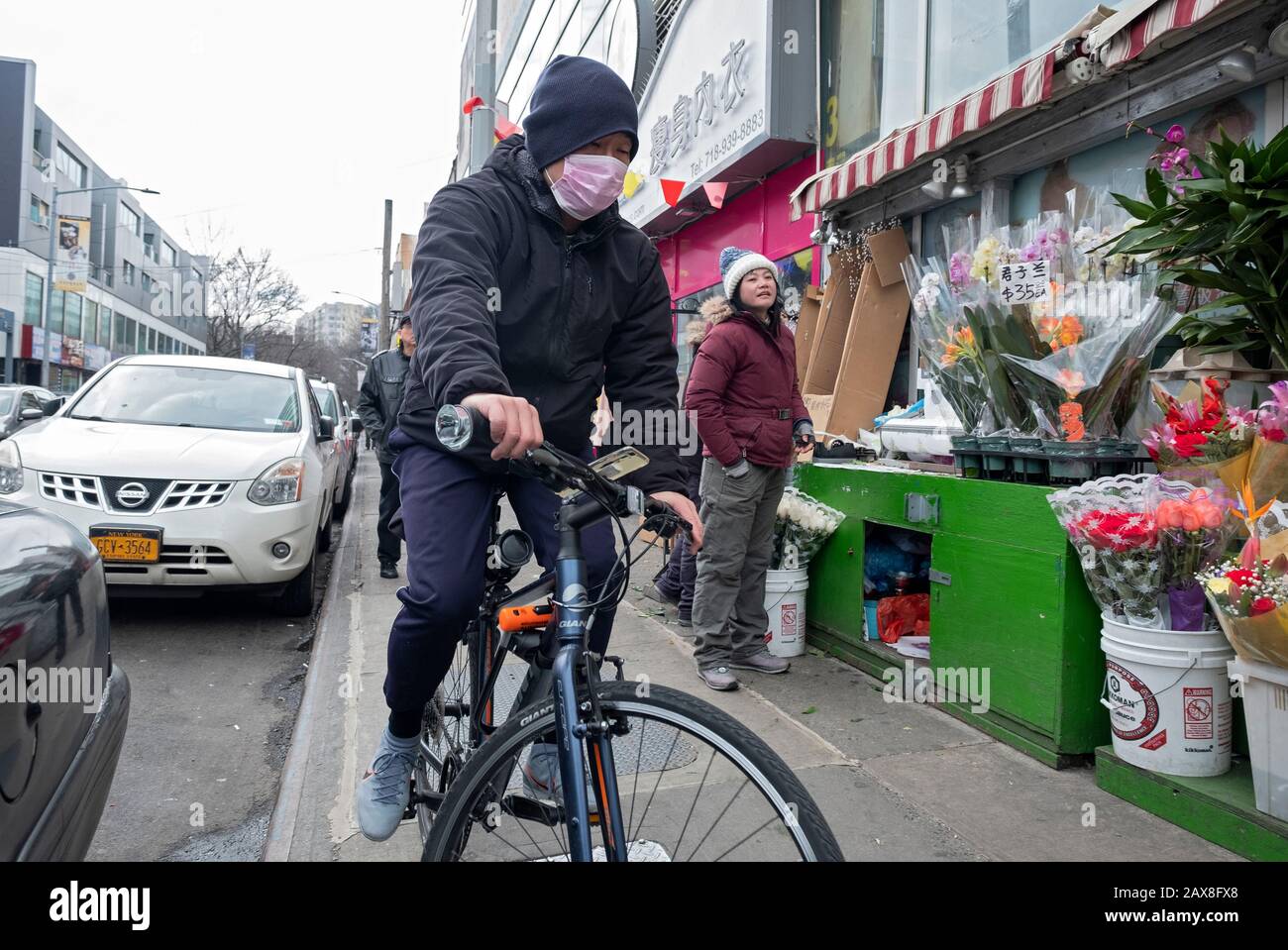 A Chinese American man wearing a surgical mask rides his bike on the sidewalk in Chinatown, Flushing, New York City. Stock Photo