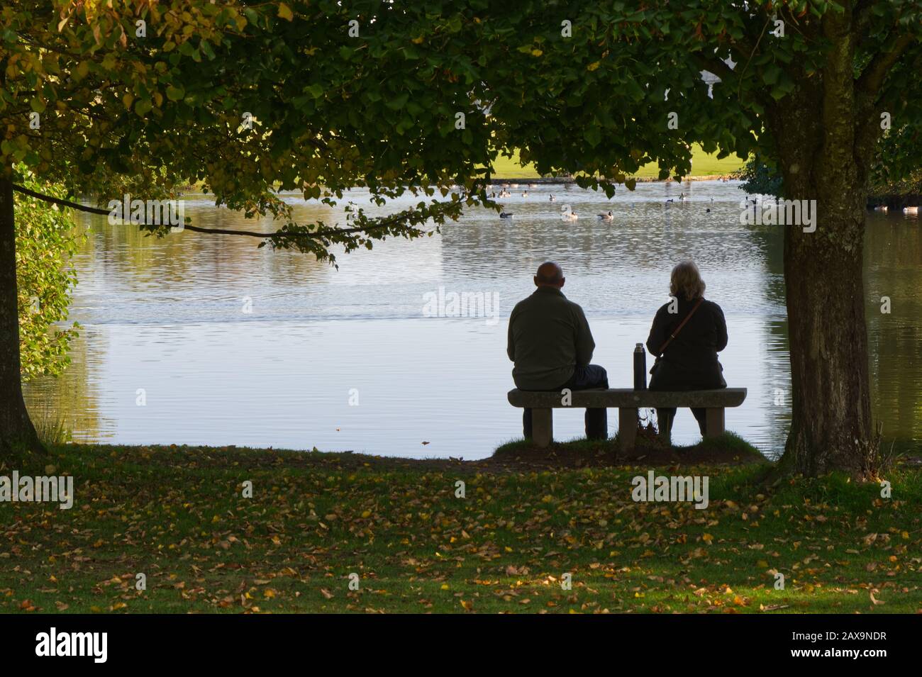 Man and woman couple sitting on seat by lake with flask. Silhouette. Stock Photo
