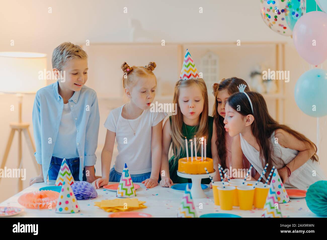 Small kids celebrate birthday party, blow candles on cake, gather at festive table, have good mood, enoy spending time together, make wish, wear party Stock Photo