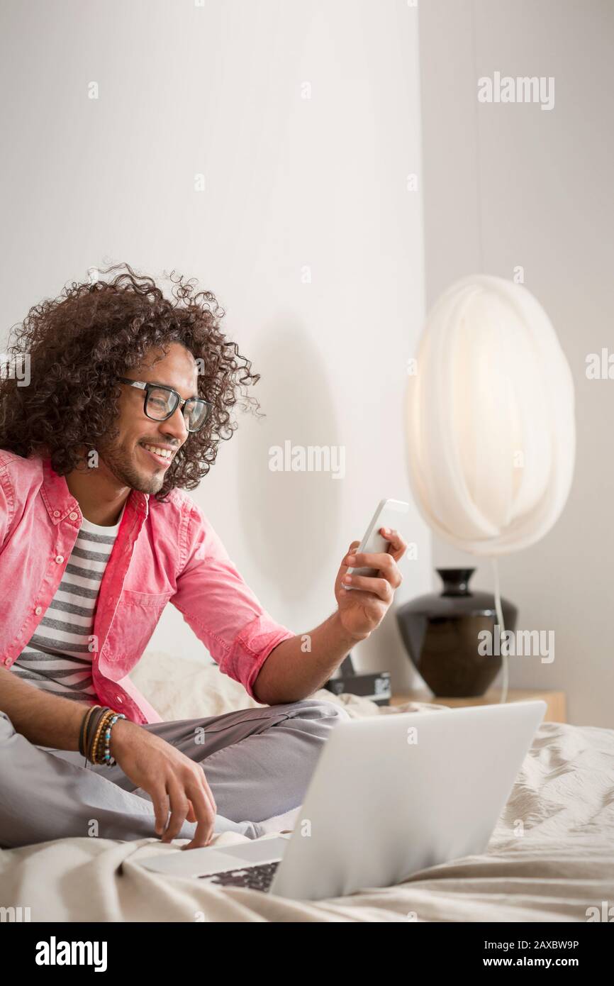Smiling young man using smart phone at laptop on bed Stock Photo