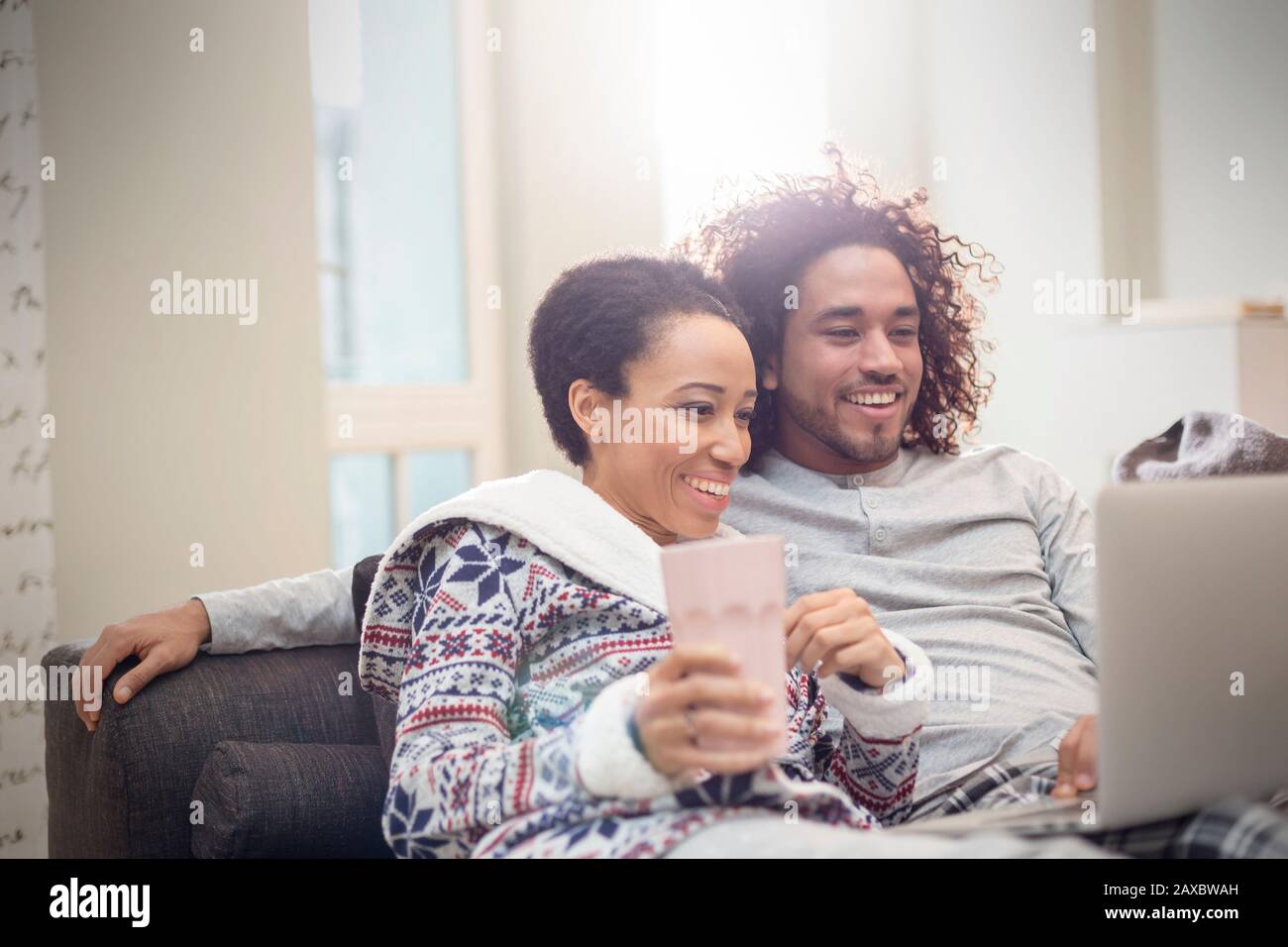 Affectionate couple in pajamas relaxing, using laptop on sofa Stock Photo