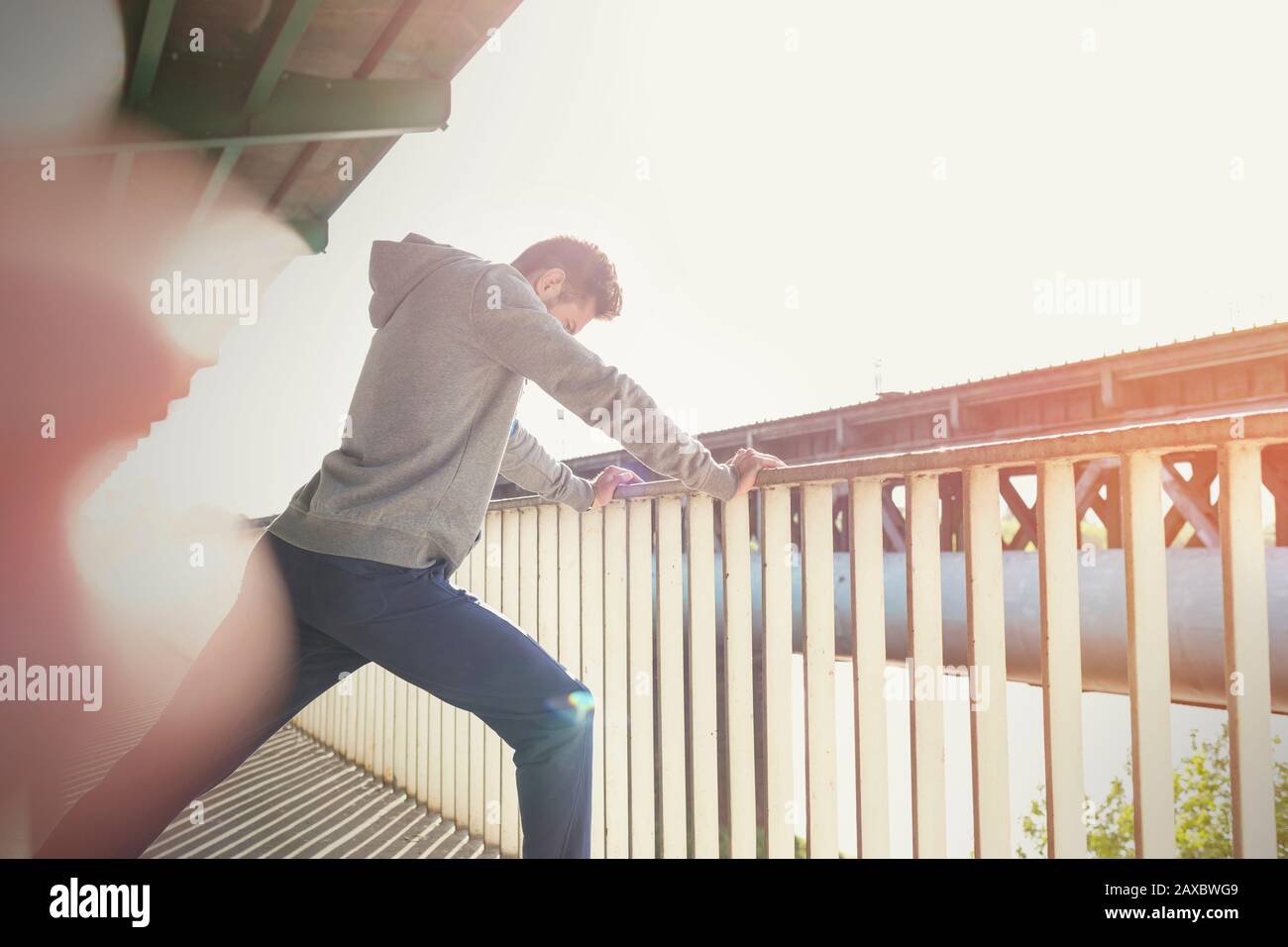 Young male runner stretching leg at sunny urban railing Stock Photo