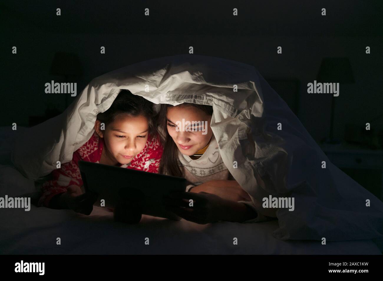 Sisters watching movie on digital tablet under blanket in dark bedroom Stock Photo