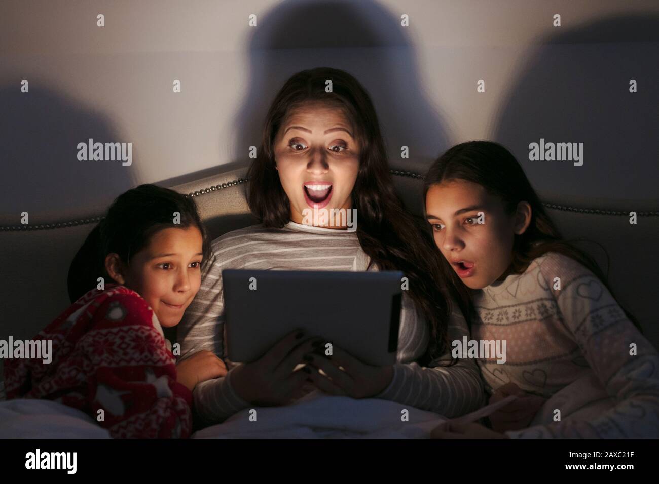 Surprised mother and daughters watching movie on digital tablet in dark bedroom Stock Photo