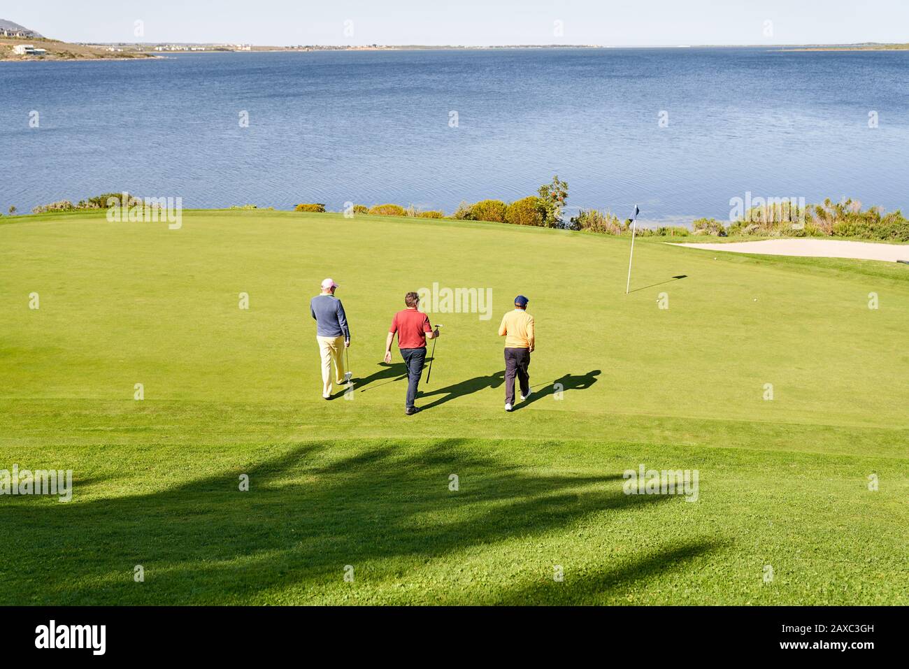 Male golfers walking toward pin on sunny lakeside putting green Stock Photo