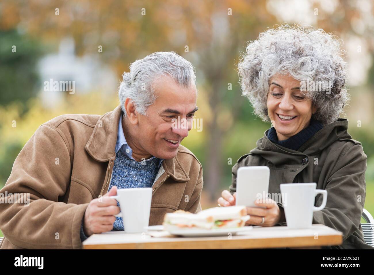 Senior couple with smart phone, eating lunch and drinking coffee in park Stock Photo