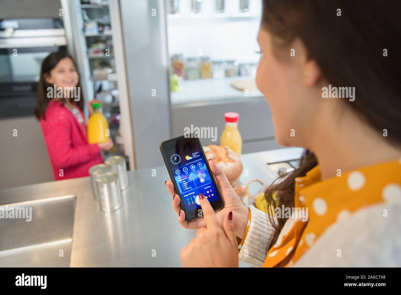Mother using smart phone app to track groceries in refrigerator, watching daughter Stock Photo