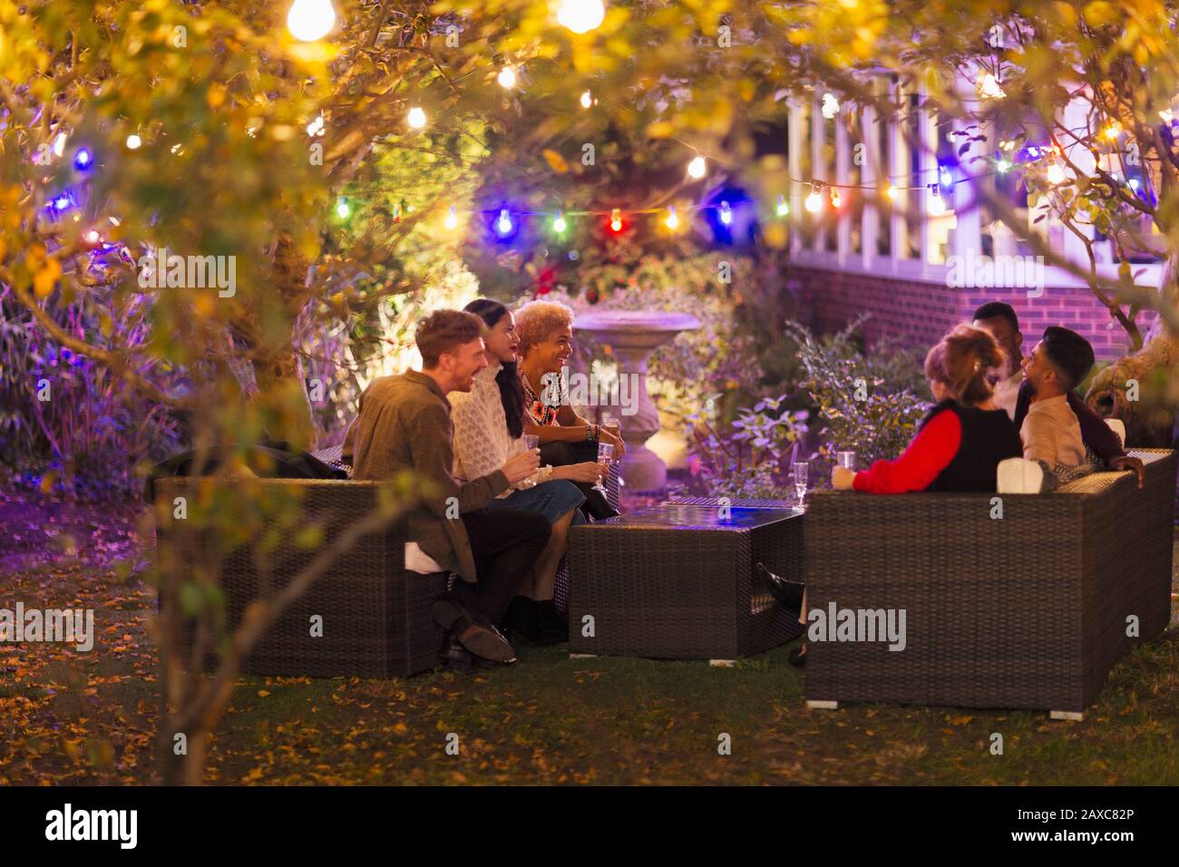 Friends talking and drinking under trees with string lights at garden party Stock Photo